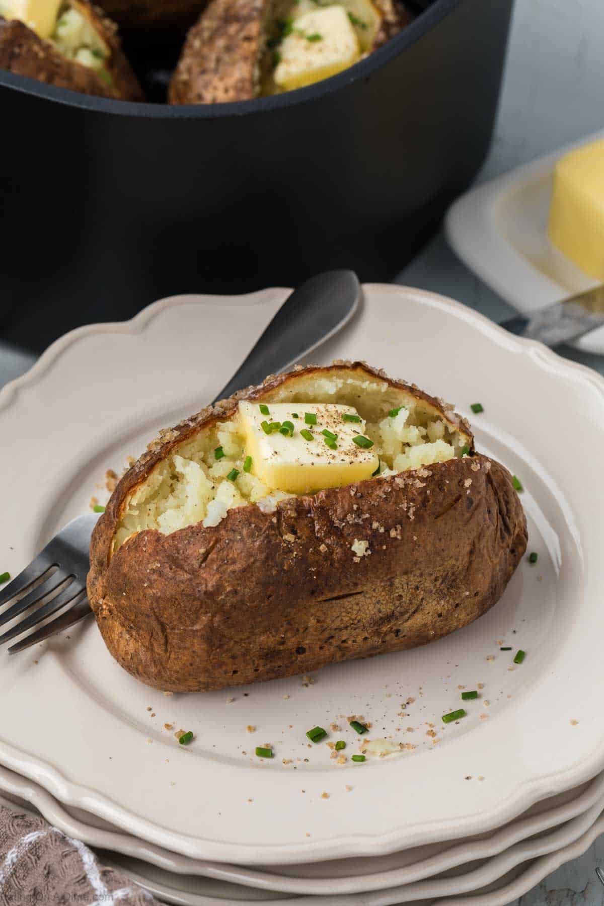 A baked potato split open on a white plate, topped with a pat of butter and sprinkled with chopped green herbs. A fork rests on the plate, and a dish of butter is visible in the background near a black container. The air fryer baked potato offers a crispy skin and fluffy interior you’ll love.
