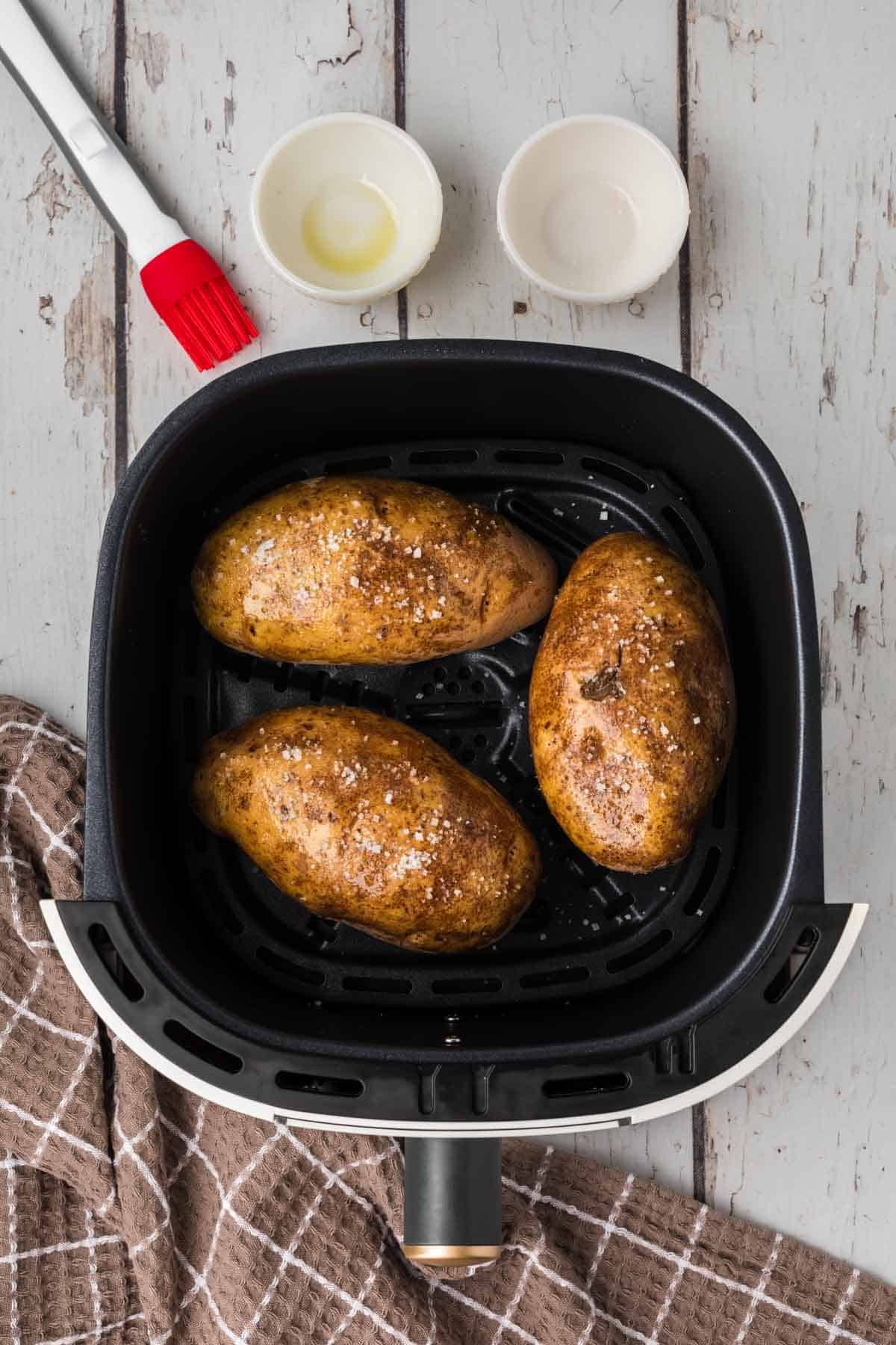 An overhead view of three air fryer baked potatoes with salt inside an air fryer basket placed on a rustic white wooden surface. A brown checkered cloth is seen in the bottom left, with a red-handled basting brush and two small bowls in the top left corner.