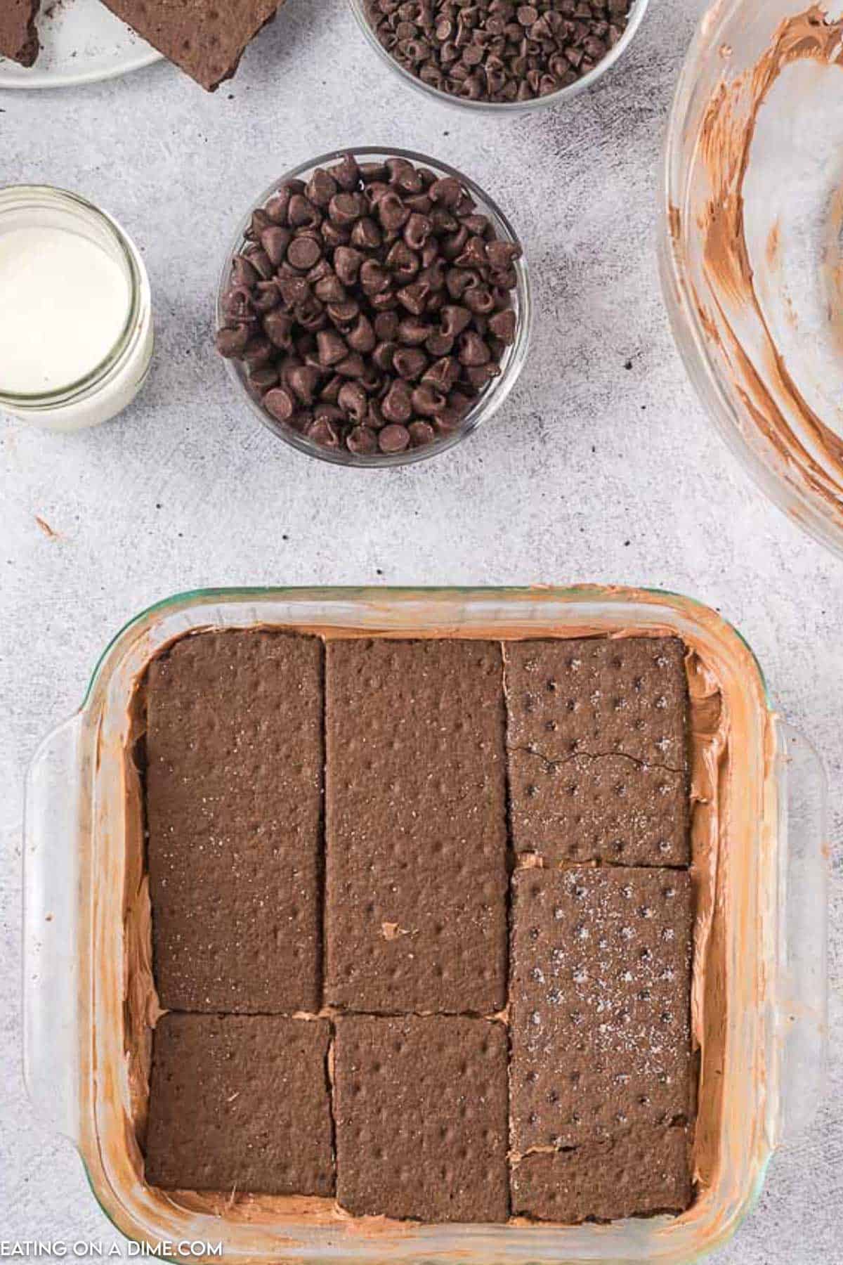 Topping the chocolate layer with a layer of chocolate graham crackers in a baking dish with small bowls of chocolate chips
