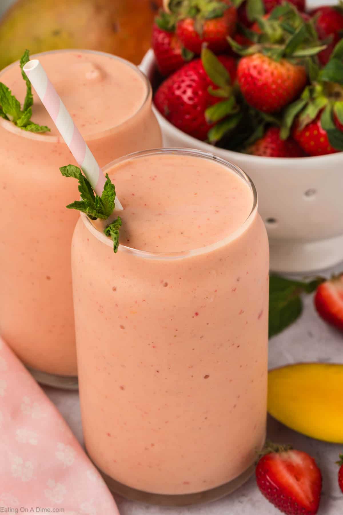 Strawberry Mango Smoothie in two glasses topped with fresh mint and a straw with a side of fresh strawberries and mango 