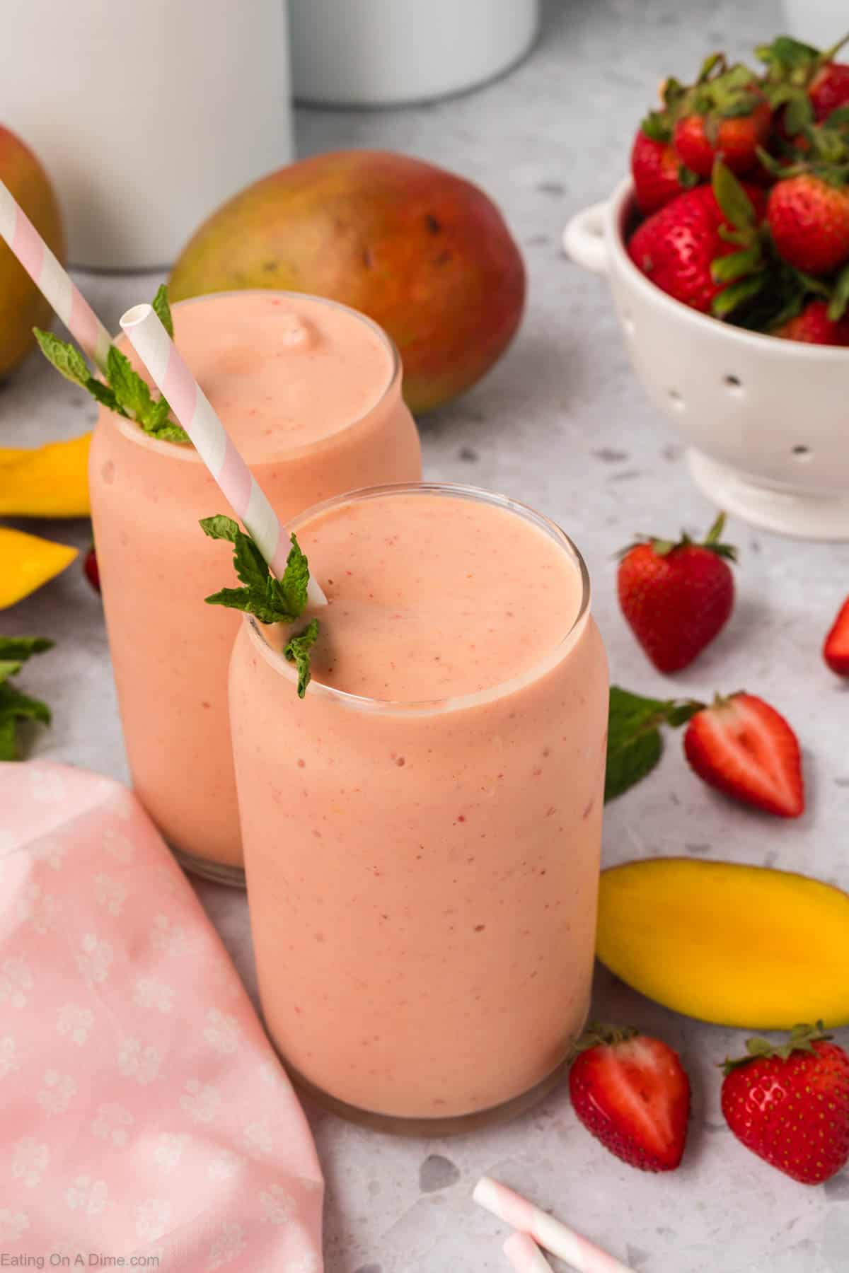Strawberry Mango Smoothie in two glasses topped with fresh mint and a straw with a side of fresh strawberries and mango