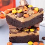 Two stacked Reese's Brownies with a thick peanut butter layer, topped with colorful peanut butter candies and pieces of peanut butter cups. Some candies are scattered on the surface around the brownies.