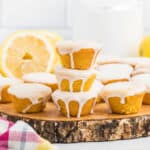 Mini Lemon Blossoms with white glaze are stacked on a rustic wooden platter. More glazed muffins and a halved lemon are in the background. A glass of milk and a colorful cloth add charm to the scene.