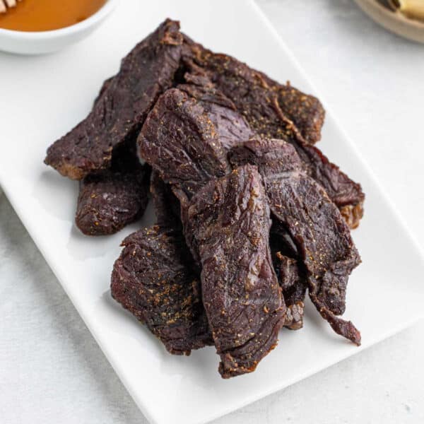 Several pieces of dark, dried Air Fryer Beef Jerky are arranged on a white rectangular plate, with a small bowl of sauce partially visible in the corner.