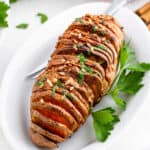 A sliced, baked Hasselback sweet potato garnished with chopped herbs and nuts, served on a white oval plate with fresh parsley—easily made in an air fryer for a crispy finish.