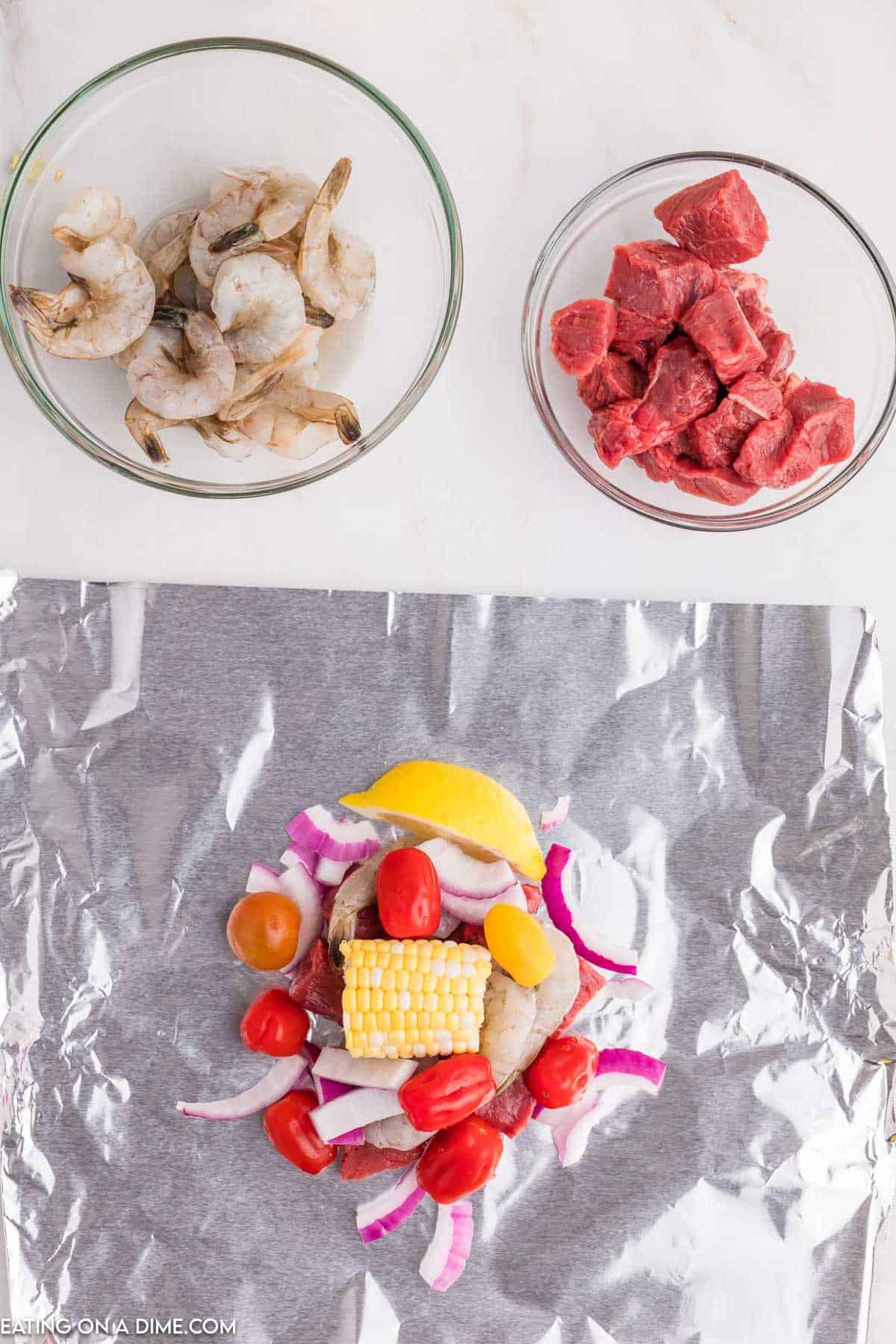 Piece of foil topped with cherry tomatoes, corn on the cob, red onion and lemon wedges with a bowl of shrimp and steak bites on the side