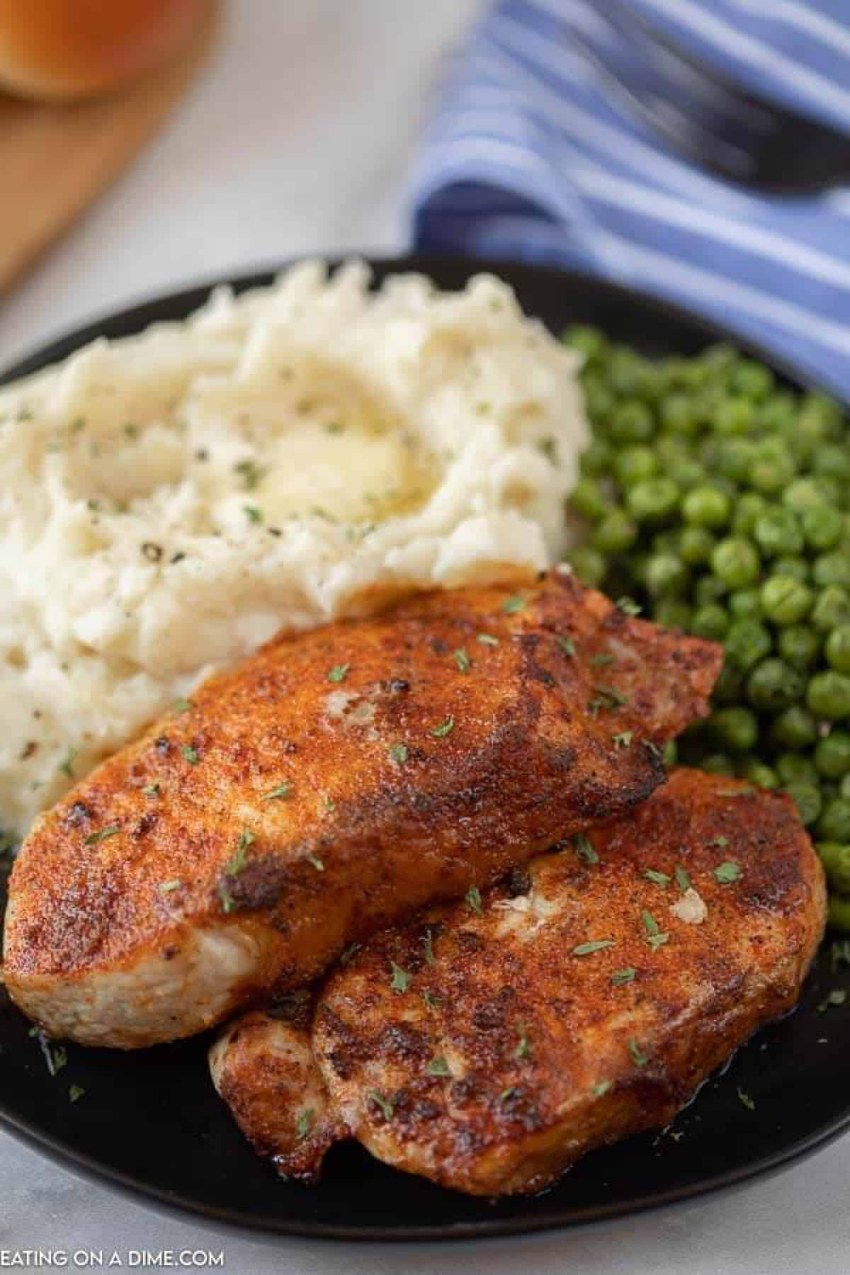Close up image of pork chops on a black plate with a side of mashed potatoes and green peas. 