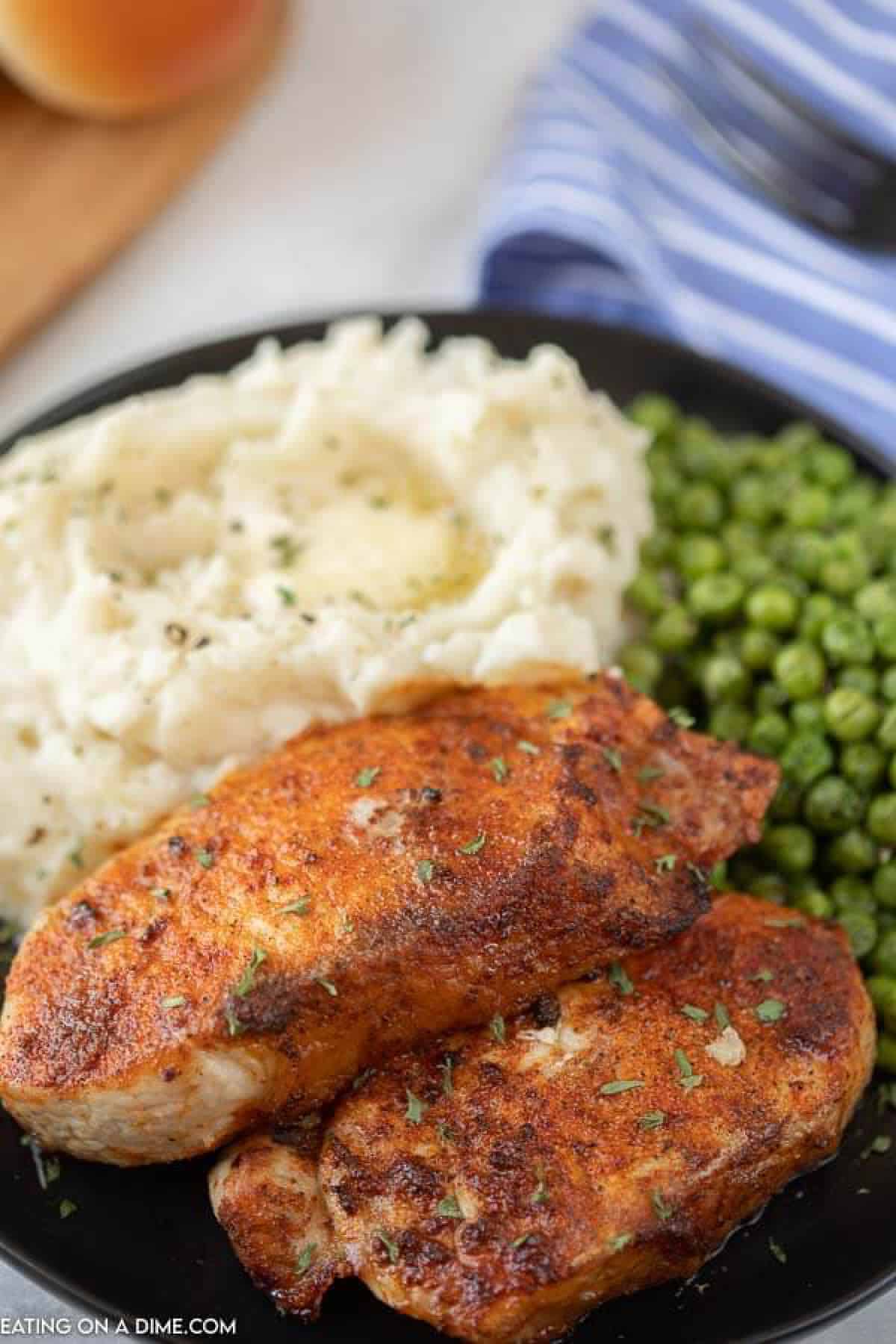 Close up image of pork chops on a black plate with a side of mashed potatoes and green peas. 