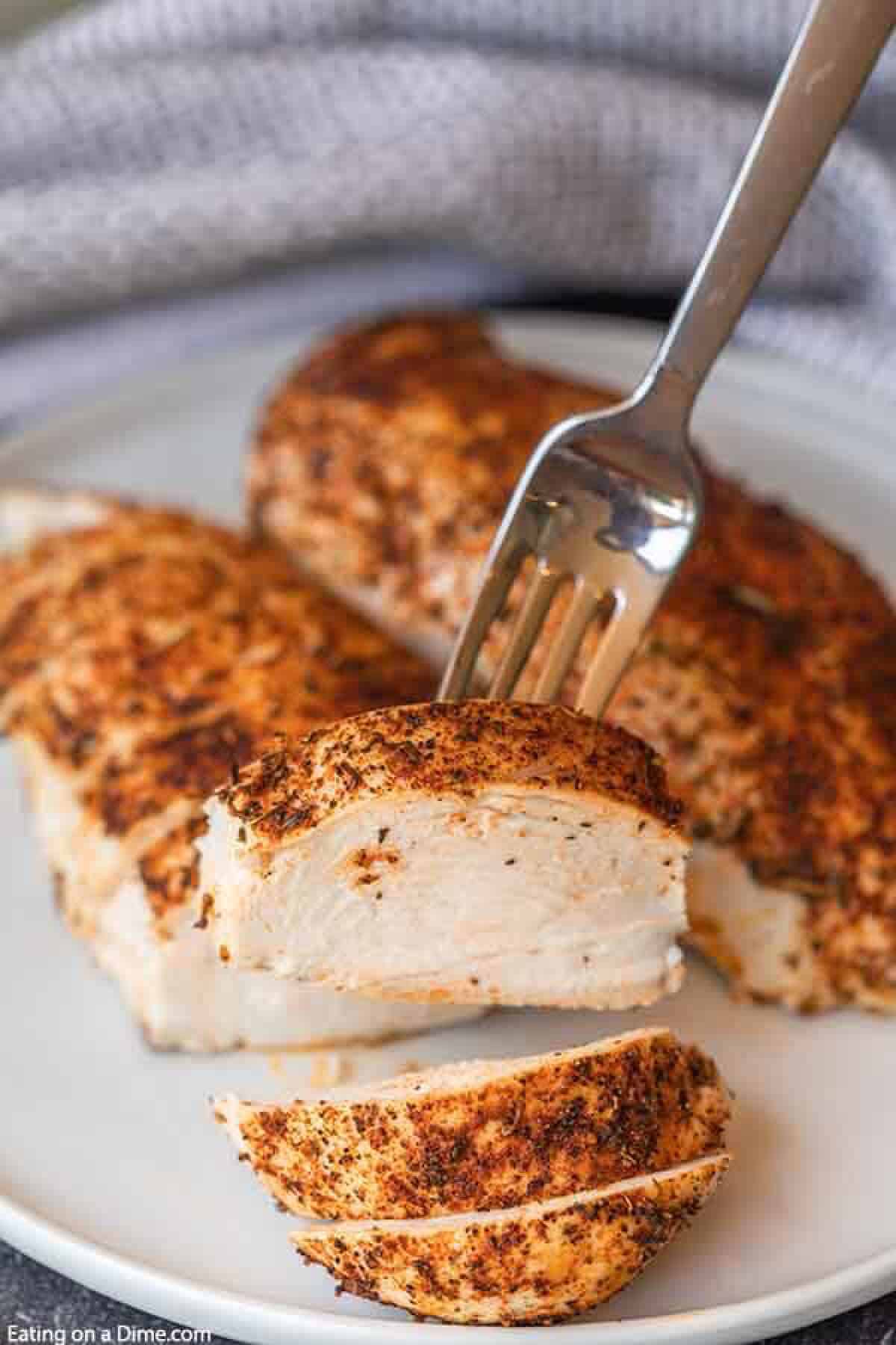 This easy baked chicken recipe has only a few ingredients and takes less than 30 minutes to bake in the oven. Learn how to bake chicken for an easy meal idea that is flavorful. Find out how to cook chicken for shredding, salads and more. How to bake chicken breast in oven. #eatingonadime #howtobakechicken #howtobakechickenintheoven #howtobakechickenintheovensimple #Intheovenhealthy #intheovenrecipes #intheovenjuicy #intheoveneasy Oven Baked Chicken on a plate sliced with a piece on a fork