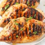 3 grilled chicken breasts on a white plate topped with fresh chopped parsely.