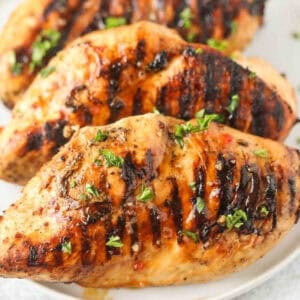 3 grilled chicken breasts on a white plate topped with fresh chopped parsely.