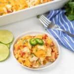A plate of cheesy nacho chicken casserole recipe with tomatoes, corn, and tortilla chips, garnished with sliced jalapeños and chopped cilantro. A blue striped napkin, fork, and lime slices are beside the plate.