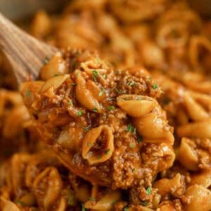 A close-up of a wooden spoon holding a serving of cheesy pasta shells mixed with ground beef in a rich tomato sauce, garnished with herbs—an irresistible beef taco pasta skillet recipe.