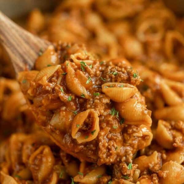 A close-up of a wooden spoon holding a serving of cheesy pasta shells mixed with ground beef in a rich tomato sauce, garnished with herbs—an irresistible beef taco pasta skillet recipe.