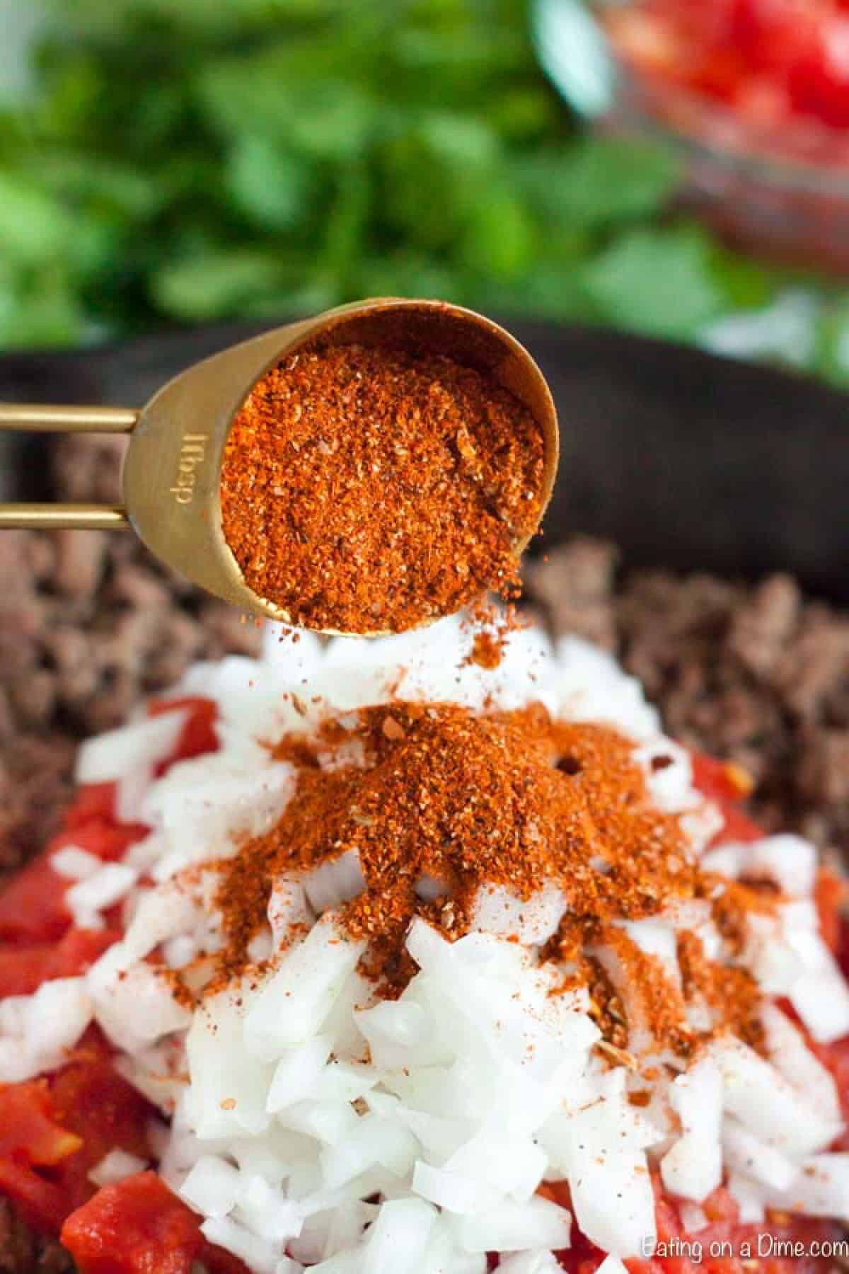 Cooked ground beef in a cast iron skillet topped with rotel, diced onions and taco seasoning being poured on top with a tablespoon