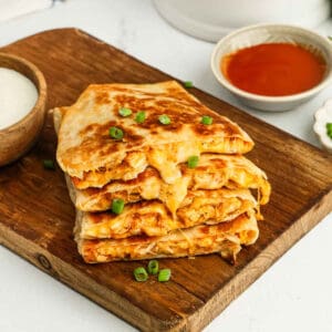 A stack of cheesy, golden-brown buffalo chicken quesadillas filled with shredded chicken and topped with chopped green onions sits on a wooden board, with bowls of dipping sauces nearby—perfect for trying out your next buffalo chicken quesadilla recipe.