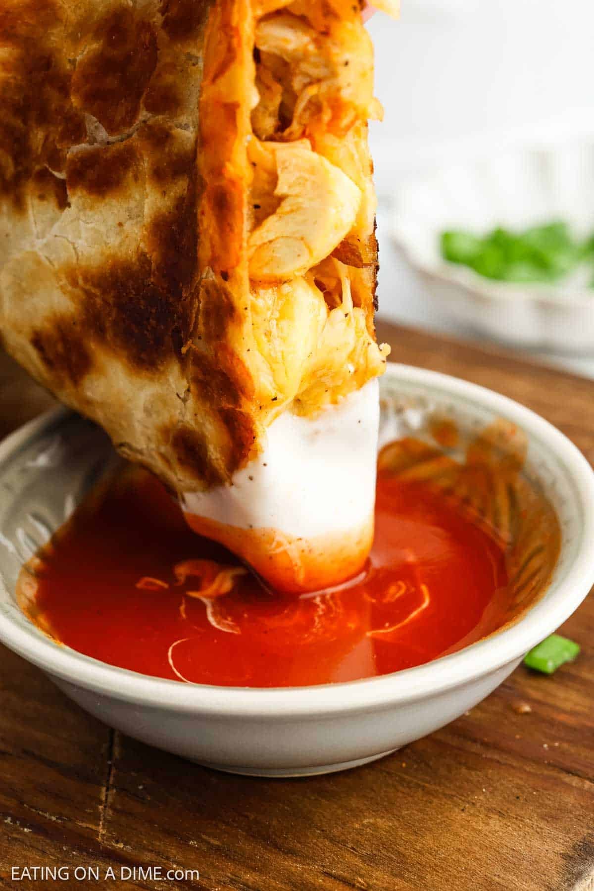 A crispy, golden burrito filled with shredded chicken is being dipped into a bowl of red sauce and white cream, reminiscent of flavors from a classic buffalo chicken quesadilla recipe, with green onions visible in the background.