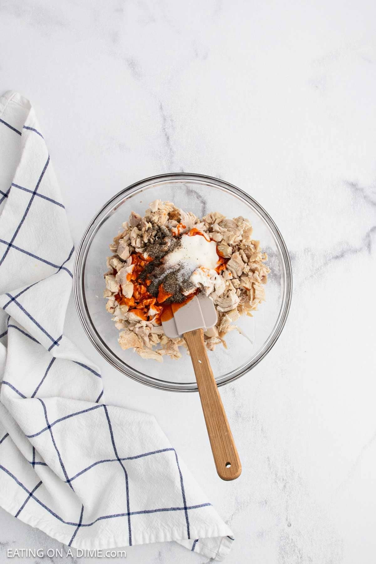 A glass bowl filled with shredded chicken, mayonnaise, spices, and seasonings sits on a white countertop beside a blue-striped towel—perfect for making a flavorful buffalo chicken quesadilla recipe. A spatula rests in the bowl for mixing.