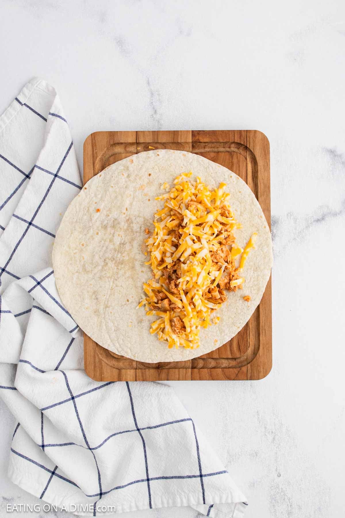 A flour tortilla filled with shredded cheese and chicken, inspired by a buffalo chicken quesadilla recipe, sits on a wooden cutting board beside a white kitchen towel with blue stripes, on a white marble surface.