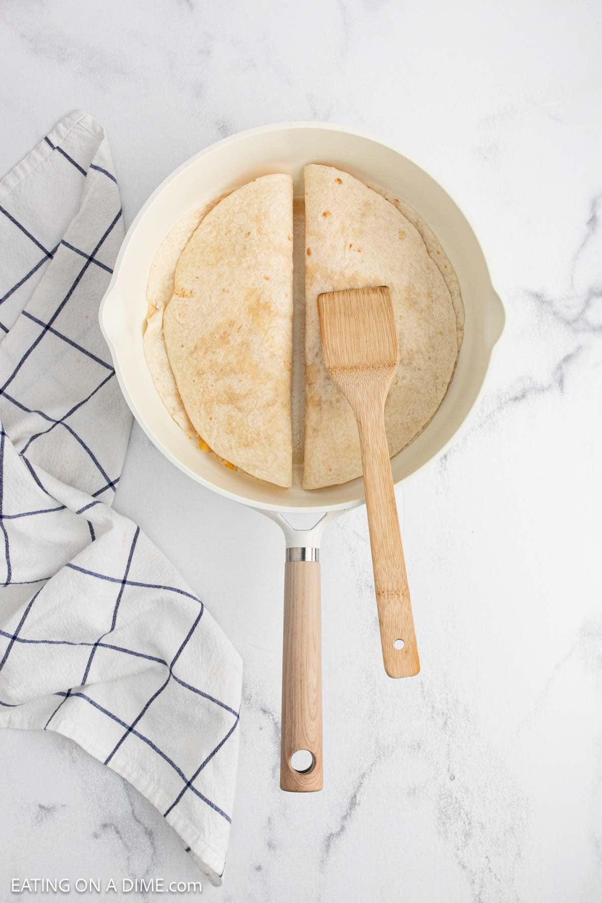 A white skillet with two folded tortillas inside, one being pressed with a wooden spatula, showcases part of a buffalo chicken quesadilla recipe. A white towel with a blue grid pattern sits beside the skillet on a white marble surface.