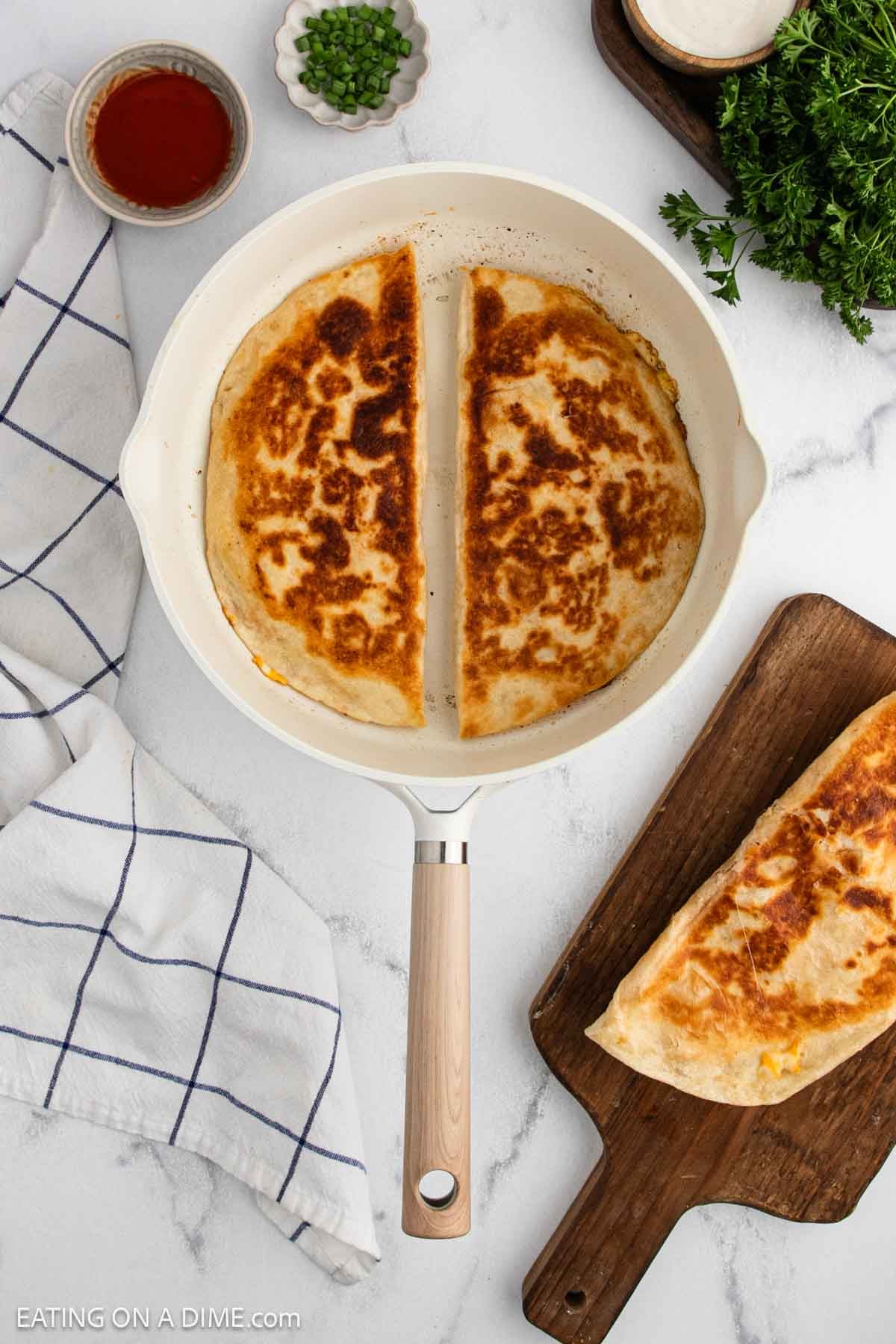 A skillet on a white surface holds two golden-brown buffalo chicken quesadilla halves. Nearby are a folded towel, a wooden board with another quesadilla, bowls of salsa, sour cream, chopped green onions, and fresh parsley.