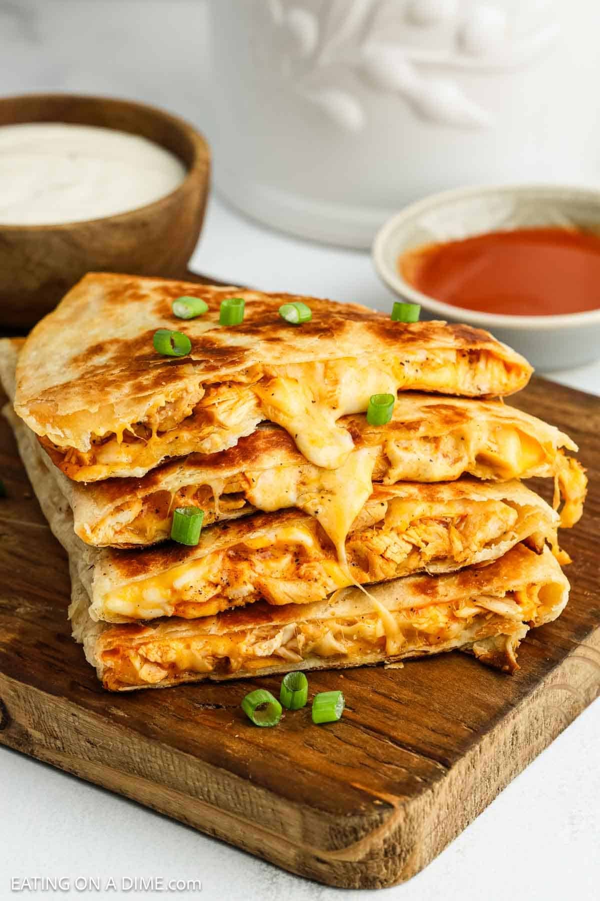 A stack of cheesy buffalo chicken quesadilla wedges sits on a wooden board. The quesadillas are garnished with chopped green onions, with small bowls of creamy dip and red sauce nearby—perfect for trying a new buffalo chicken quesadilla recipe.