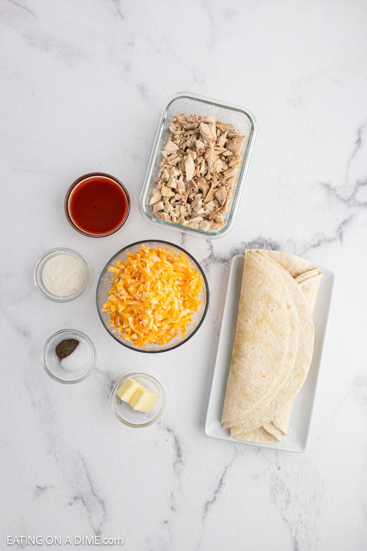 A flat lay of ingredients on a white surface for a buffalo chicken quesadilla recipe: shredded chicken, shredded cheese, tortillas, red sauce, white sauce, butter, salt, and pepper.