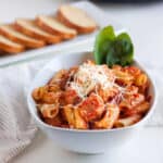A white bowl filled with Crockpot Pasta and Meatballs in tomato sauce, topped with grated cheese and fresh basil leaves. Slices of bread are on a plate in the background.
