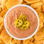 A white bowl of creamy Copycat Fritos Bean Dip Recipe topped with sliced jalapeños, surrounded by corn chips.