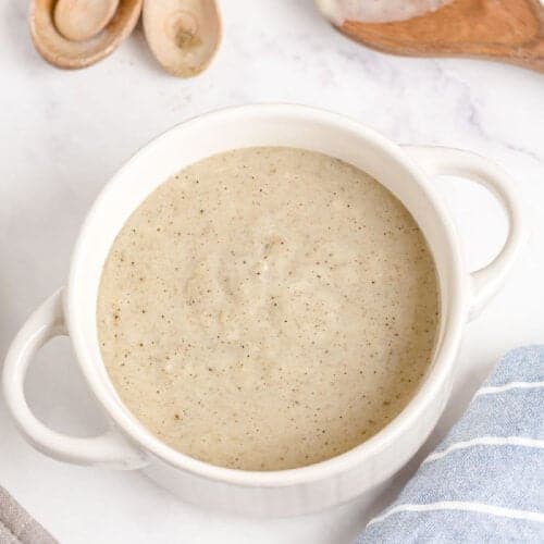 A white bowl filled with creamy, light beige Homemade Cream of Chicken Soup sits on a white surface, with wooden spoons and a blue striped cloth nearby.