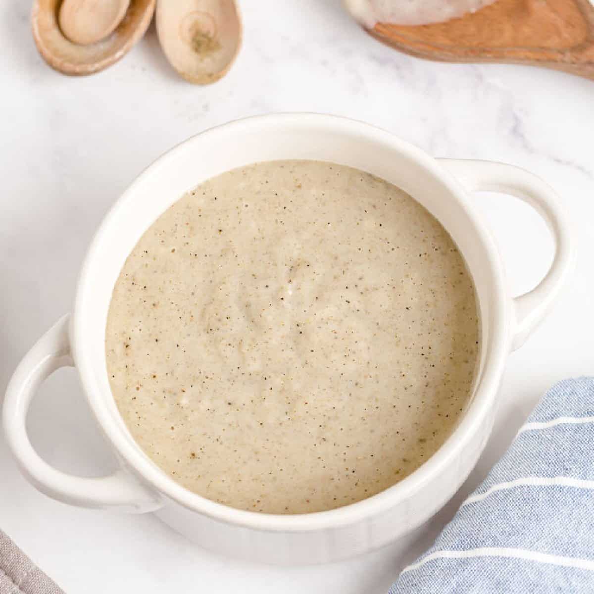 A white bowl filled with creamy, light beige Homemade Cream of Chicken Soup sits on a white surface, with wooden spoons and a blue striped cloth nearby.