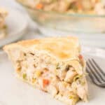 A close-up of a slice of creamy Double Crust Chicken Pot Pie with vegetables in a flaky crust, served on a white plate next to a fork. The rest of the homemade pie is visible in the background.