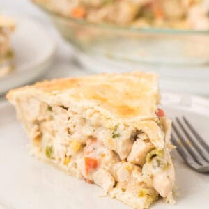 A close-up of a slice of creamy Double Crust Chicken Pot Pie with vegetables in a flaky crust, served on a white plate next to a fork. The rest of the homemade pie is visible in the background.