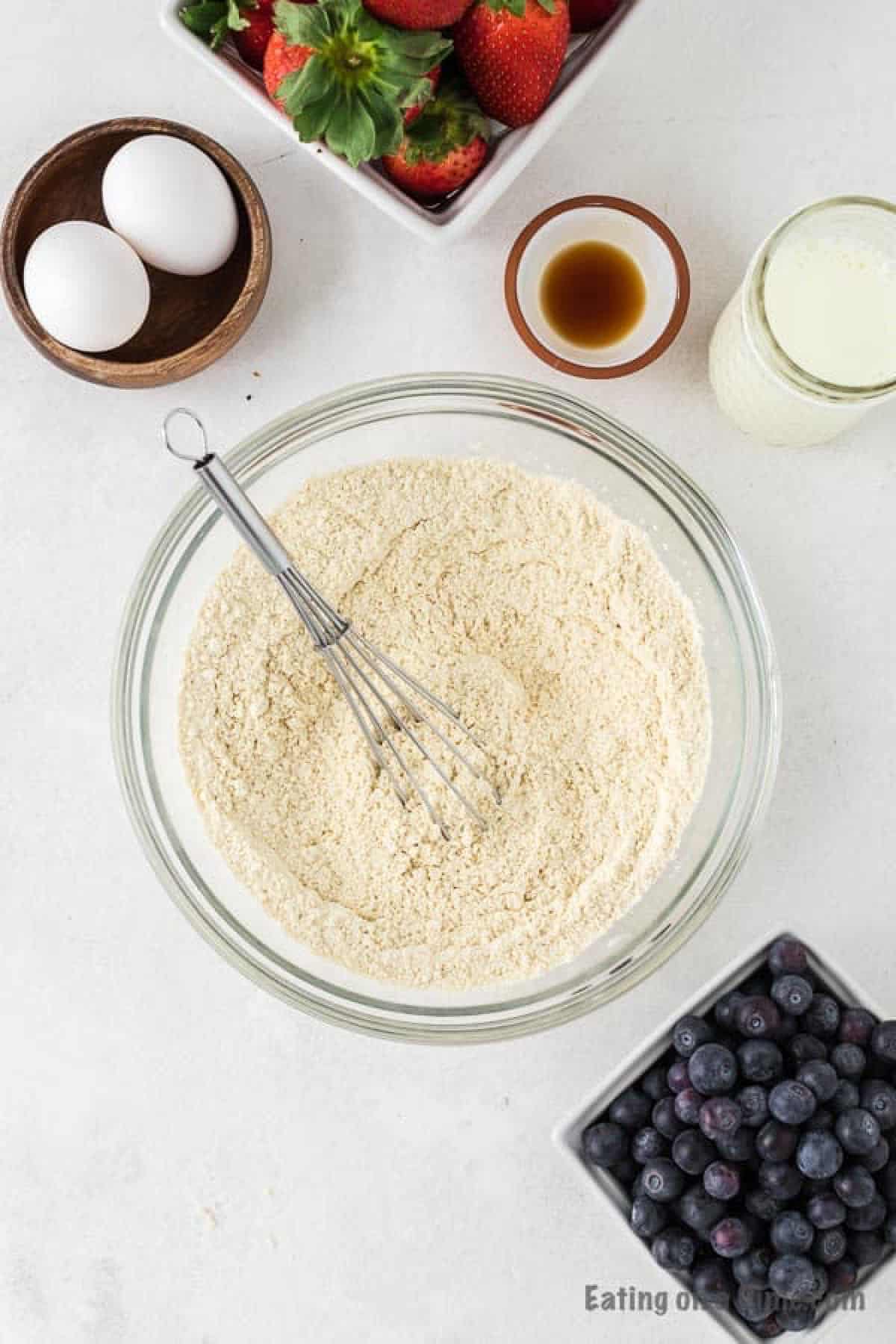 Combining the dry ingredients in a bowl with a whisk with a bowl of eggs, vanilla extract, milk and blueberries on the side