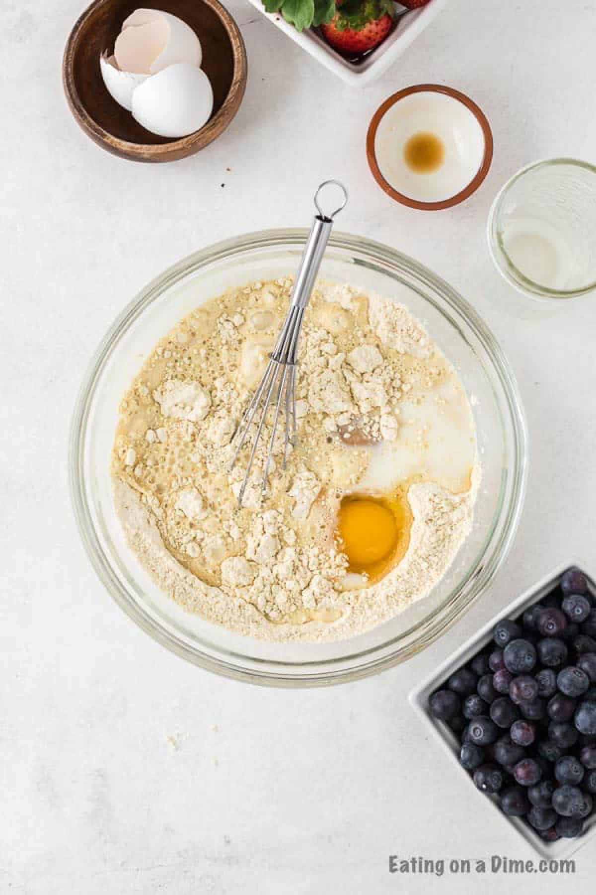 Adding the milk and eggs to the flour mixture with a bowl of cracked eggs and fresh blueberries on the side
