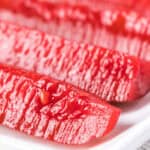 Close-up of several slices of bright red, Kool Aid Pickles pickled watermelon rind arranged on a white surface. The slices show a glossy texture and visible seeds.