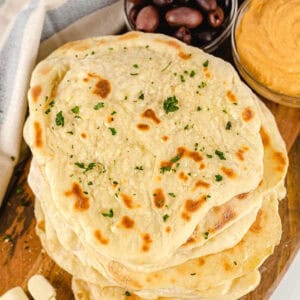 A stack of freshly cooked naan bread, made using a simple How to Make Naan Bread recipe, is garnished with chopped herbs and served on a wooden board alongside black olives and orange hummus.