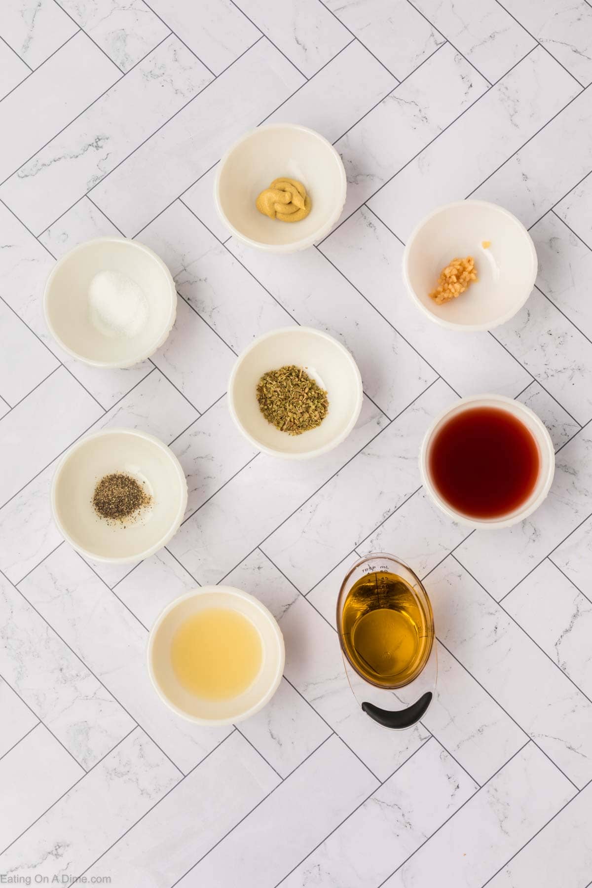 Seven small bowls with various ingredients and two small containers with liquids are arranged on a white patterned surface. The ingredients, perfect for making an orzo pasta salad, appear to include spices, herbs, and condiments used for cooking or preparing a dish.