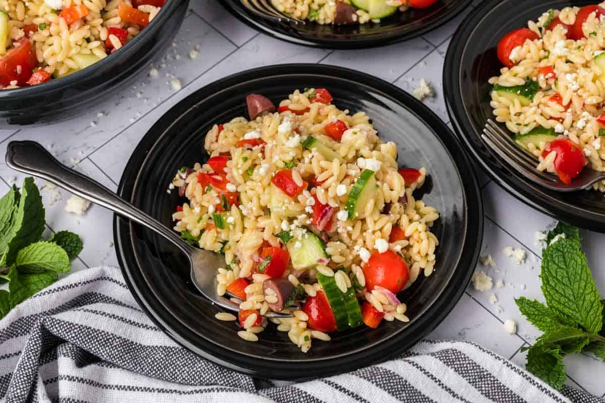 A black plate filled with a colorful orzo pasta salad, featuring cherry tomatoes, cucumber slices, red bell pepper, feta cheese, and bits of red onion. A fork rests on the plate, and a striped cloth napkin is placed nearby. Additional plates of salad are visible in the background.