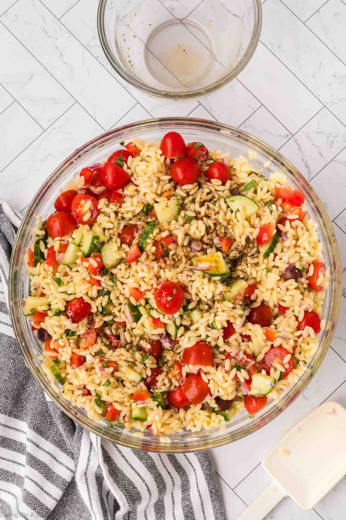 A bowl of fresh orzo salad filled with cherry tomatoes, cucumbers, olives, and herbs is placed on a white tiled surface. A dirty mixing bowl is in the background, and a striped kitchen towel is at the bottom left corner of the image.