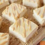 Squares of creamy Peanut Butter Cheesecake Bites with a crumbly crust, topped with light caramel or butterscotch drizzle, are arranged on a wooden board. The bars have smooth, pale surfaces with a decorative topping.