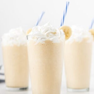 Four glasses of Peanut Butter Banana Smoothie topped with whipped cream, garnished with banana slices and blue-striped straws, set against a white background.