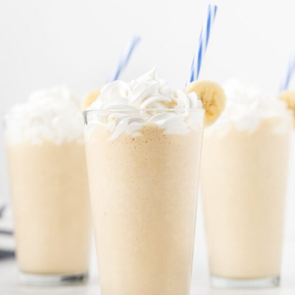 Four glasses of Peanut Butter Banana Smoothie topped with whipped cream, garnished with banana slices and blue-striped straws, set against a white background.