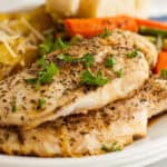 Two seasoned, baked Balsamic Sheet Pan Chicken breasts garnished with chopped parsley, served on a white plate with roasted vegetables like carrots and potatoes in the background.