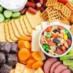 A colorful summer snack charcuterie board with crackers, pretzels, cheese slices, dried apricots, chocolate pieces, sliced meats, fresh cucumber, a dip, and a bowl of mixed nuts, raisins, and candy-coated chocolates.
