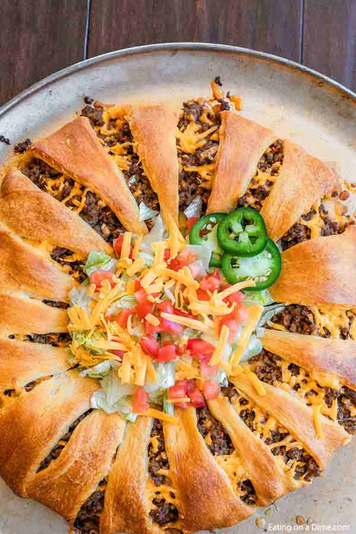 Taco Crescent Roll Ring topped with lettuce, tomato, shredded cheese and jalapeños on a pizza pan. 