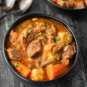 A bowl of hearty Crock Pot Beef Stew Recipe with chunks of beef, carrots, potatoes, green beans, and a rich broth, garnished with chopped parsley. Spoons are visible in the background.