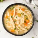 A bowl of Crock Pot chicken noodle soup with shredded chicken, sliced carrots, celery, and fresh parsley in a light broth, viewed from above on a white surface.