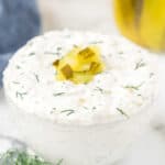 A small glass bowl filled with creamy dip garnished with chopped pickles and fresh dill on top, showcasing a delicious dill pickle dip recipe on a white surface.