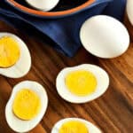 Several instant pot hard boiled eggs, some peeled and halved to reveal their yellow yolks, are arranged on a wooden surface next to an unpeeled egg and a dark blue napkin.